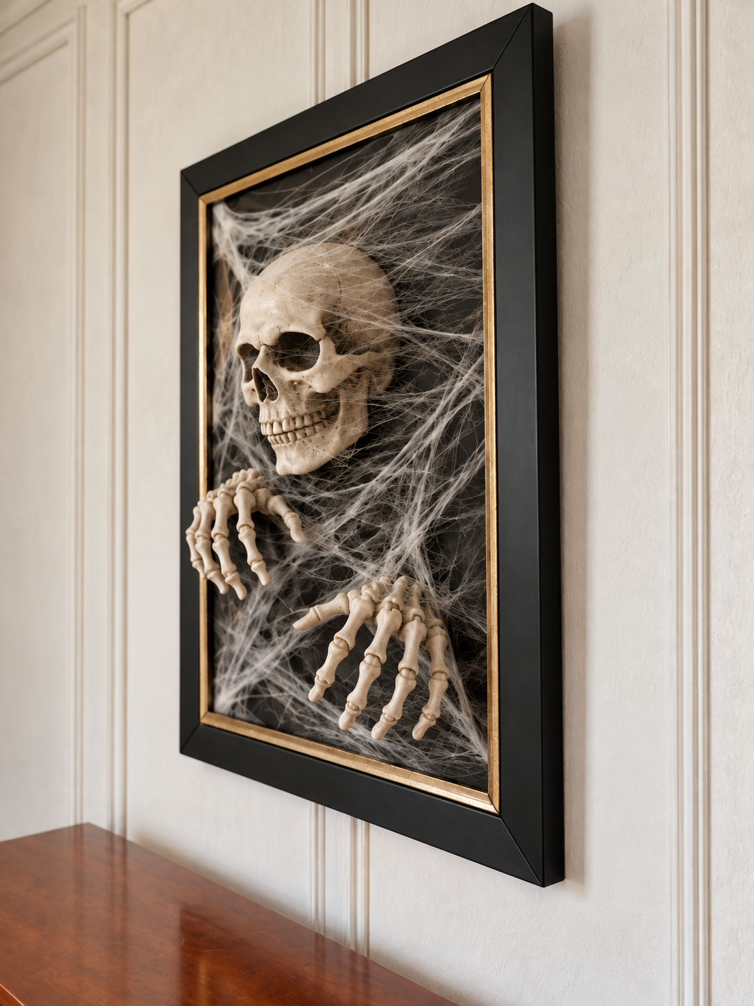 Finished skeleton wall art with a skull, two reaching hands, layered cobwebs, and a smooth black-and-gold frame photographed from beside the buffet